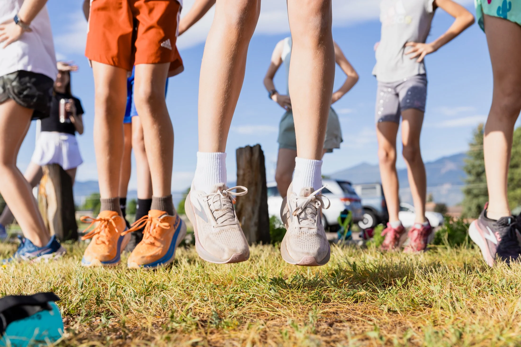 A group of people standing outdoors with their legs visible from the knees down, wearing athletic shoes, on a grassy field with mountains and a cloudy sky in the background.