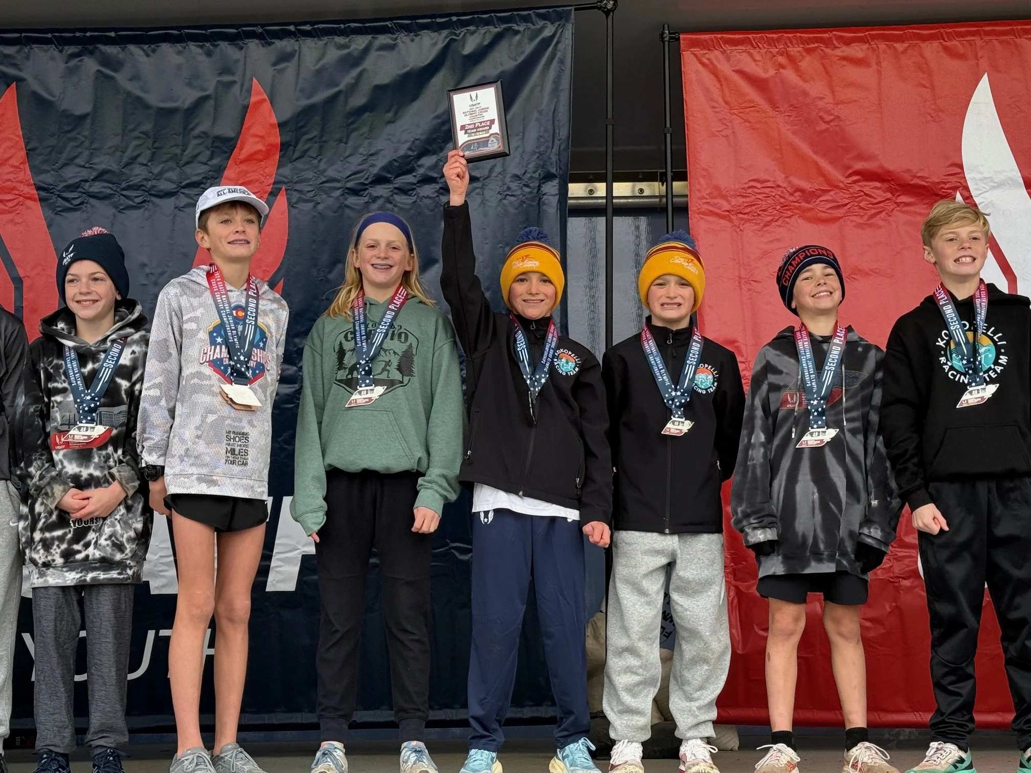 Group of young athletes celebrating on podium with medals, holding a framed award, and standing in front of black, navy, and red banners.