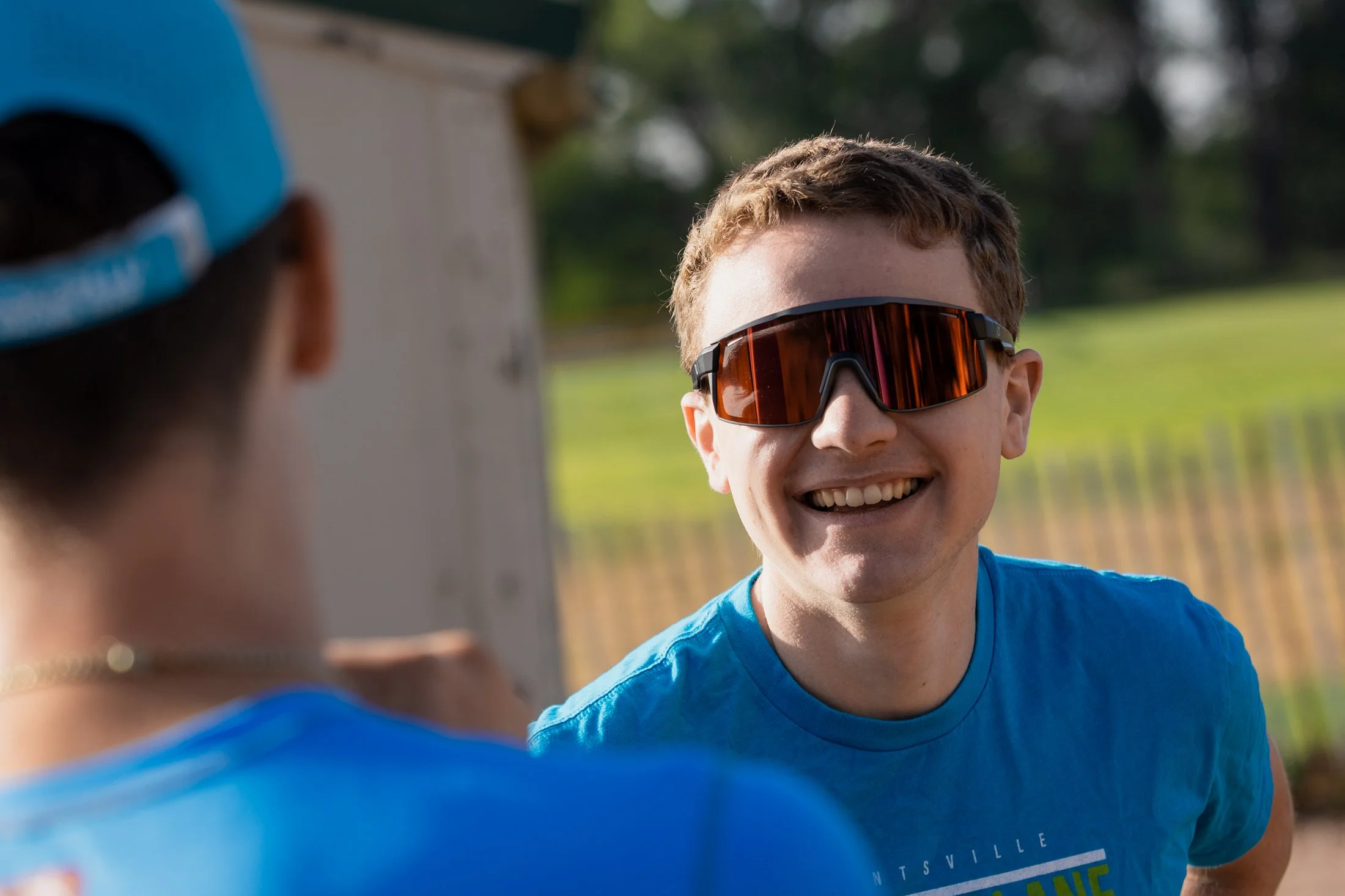Young smiling man wearing reflective sports sunglasses and a blue T-shirt outdoors, with blurred background of trees and a person in the foreground wearing a blue cap and shirt.