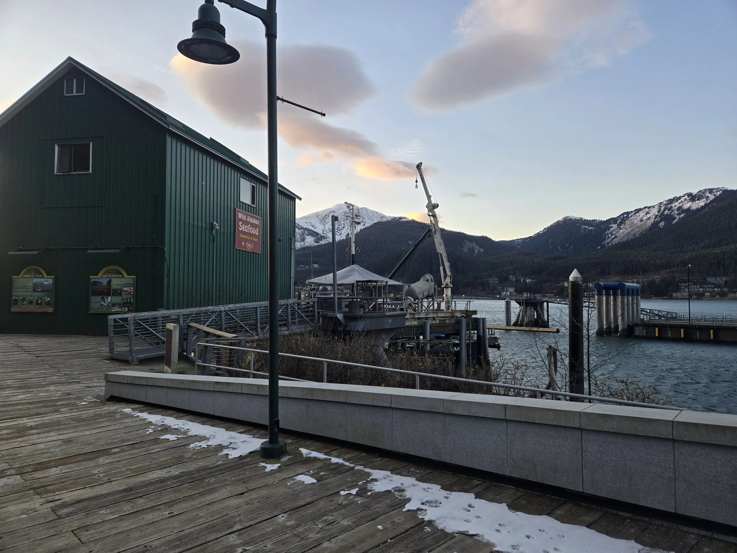 A lakeside dock with a green building, crane, boat, and mountains in the background during the evening with cloudy sky.
