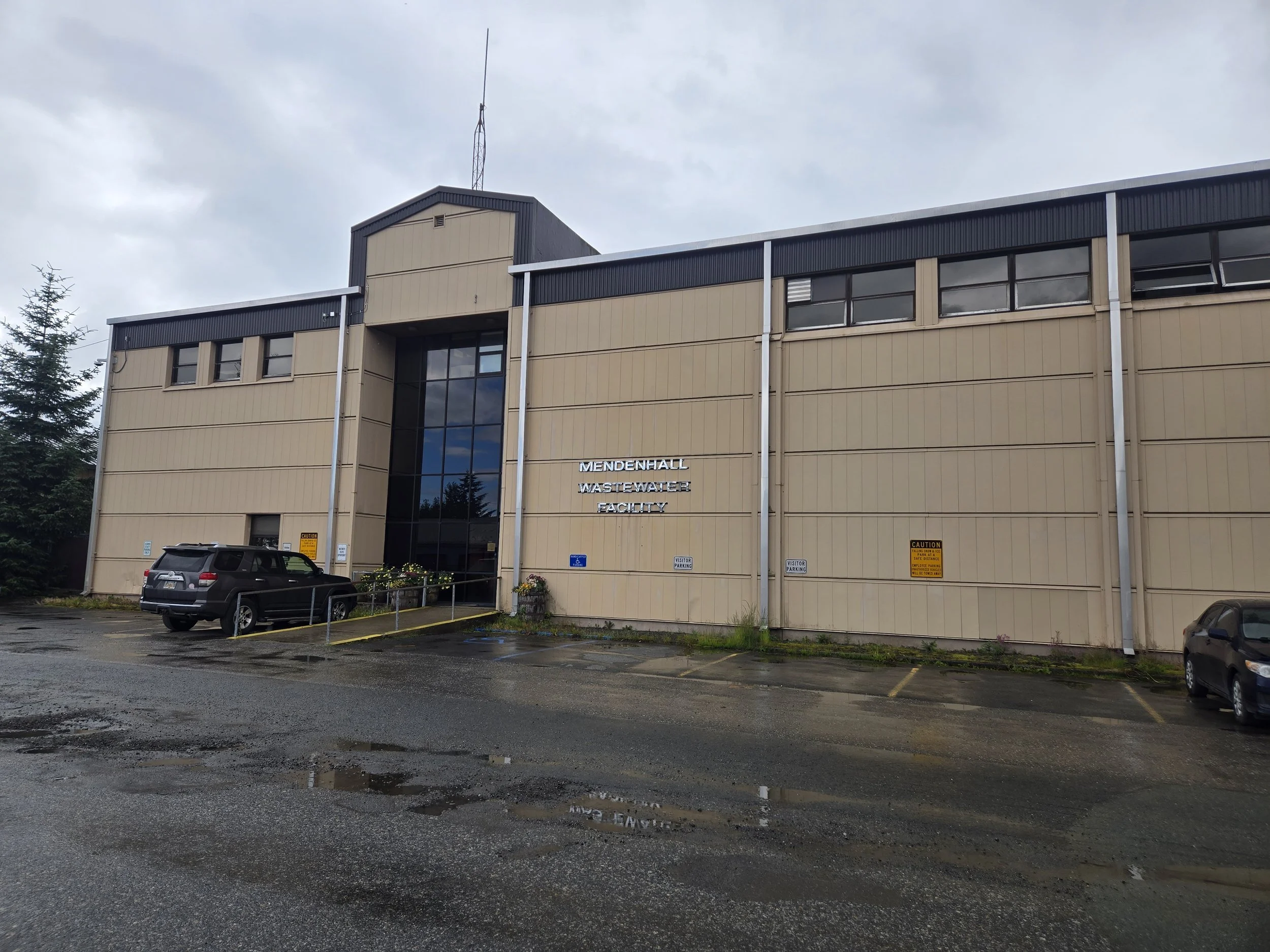 The image shows the exterior of the Mendenhall Wastewater Facility, a multi-story building with beige panels and black window frames, with a parking lot in front that has a few cars and puddles of water on the ground. There are signs indicating visitor parking, and a cloudy sky above.