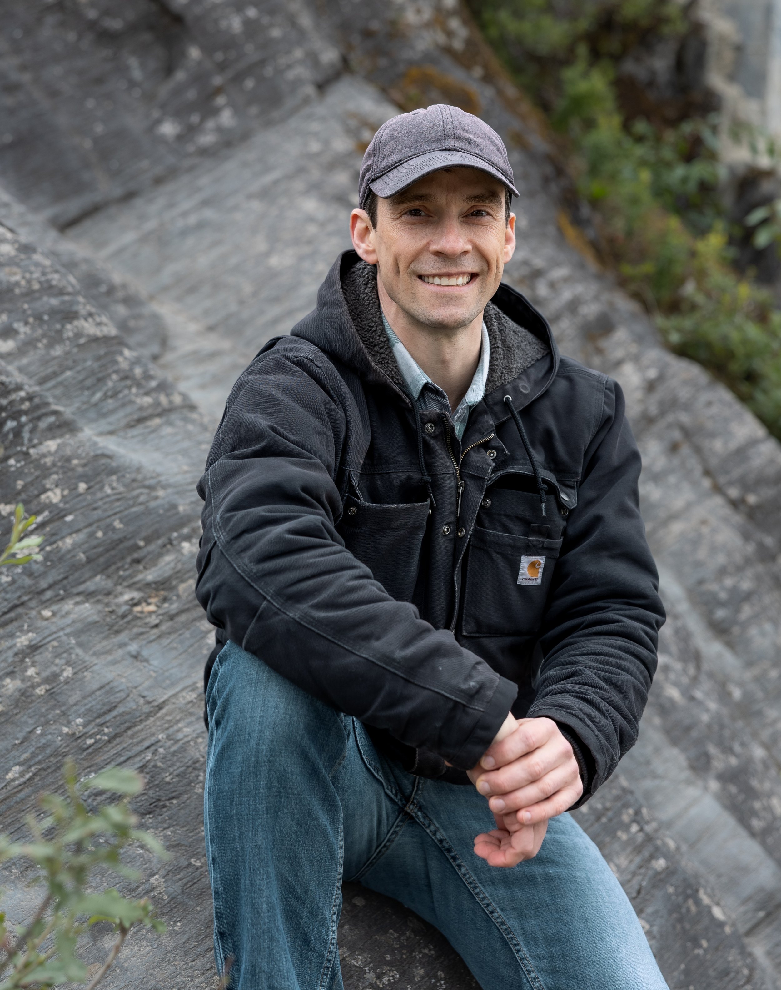 Malachi sitting on a rock, smiling and wearing a gray cap, a dark jacket, and jeans, with a background of rocks and green foliage.
