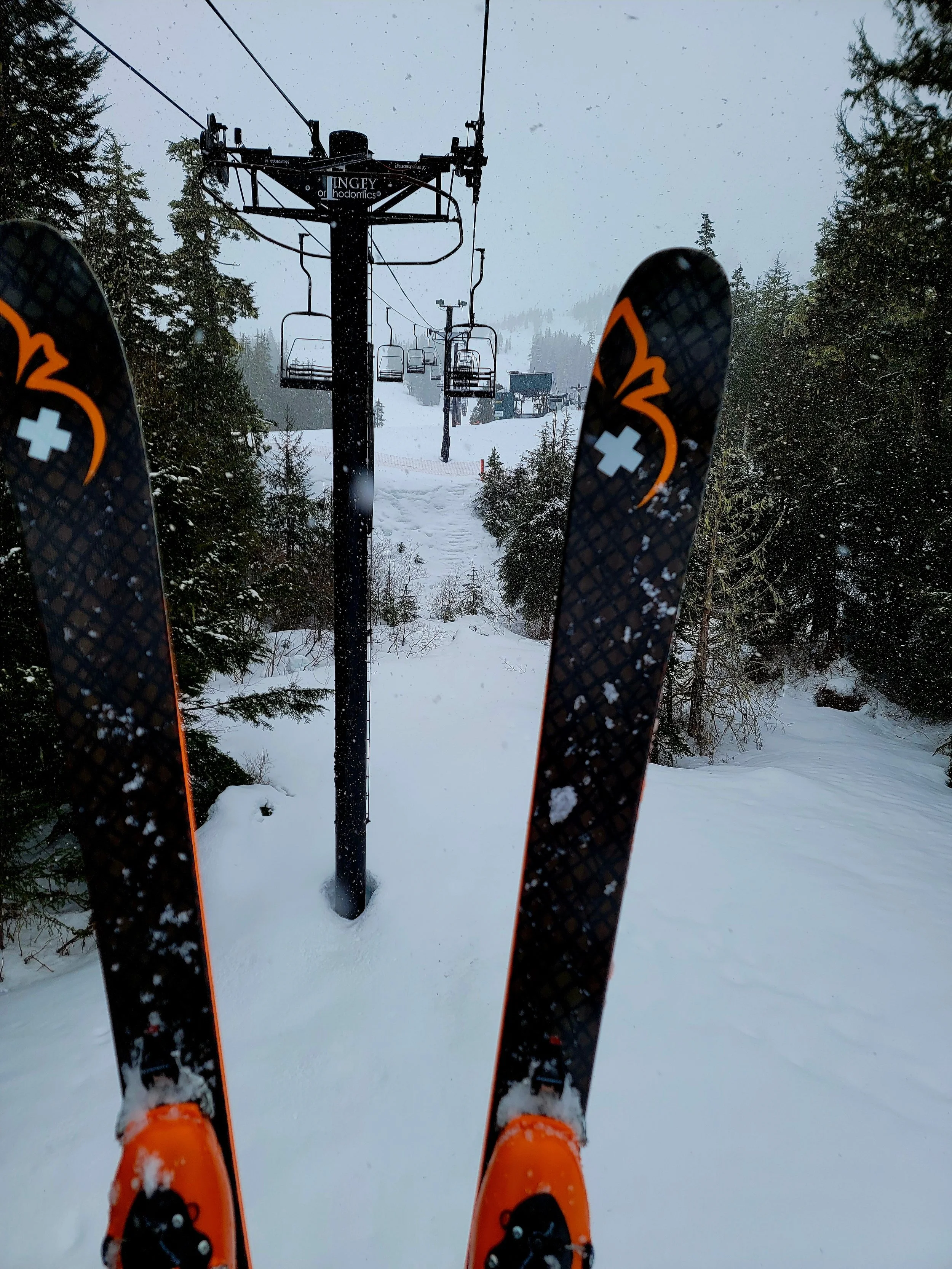 First-person view of a skier wearing orange ski boots and black skis, looking at ski lift chairs and snowy landscape in a forested mountain area.