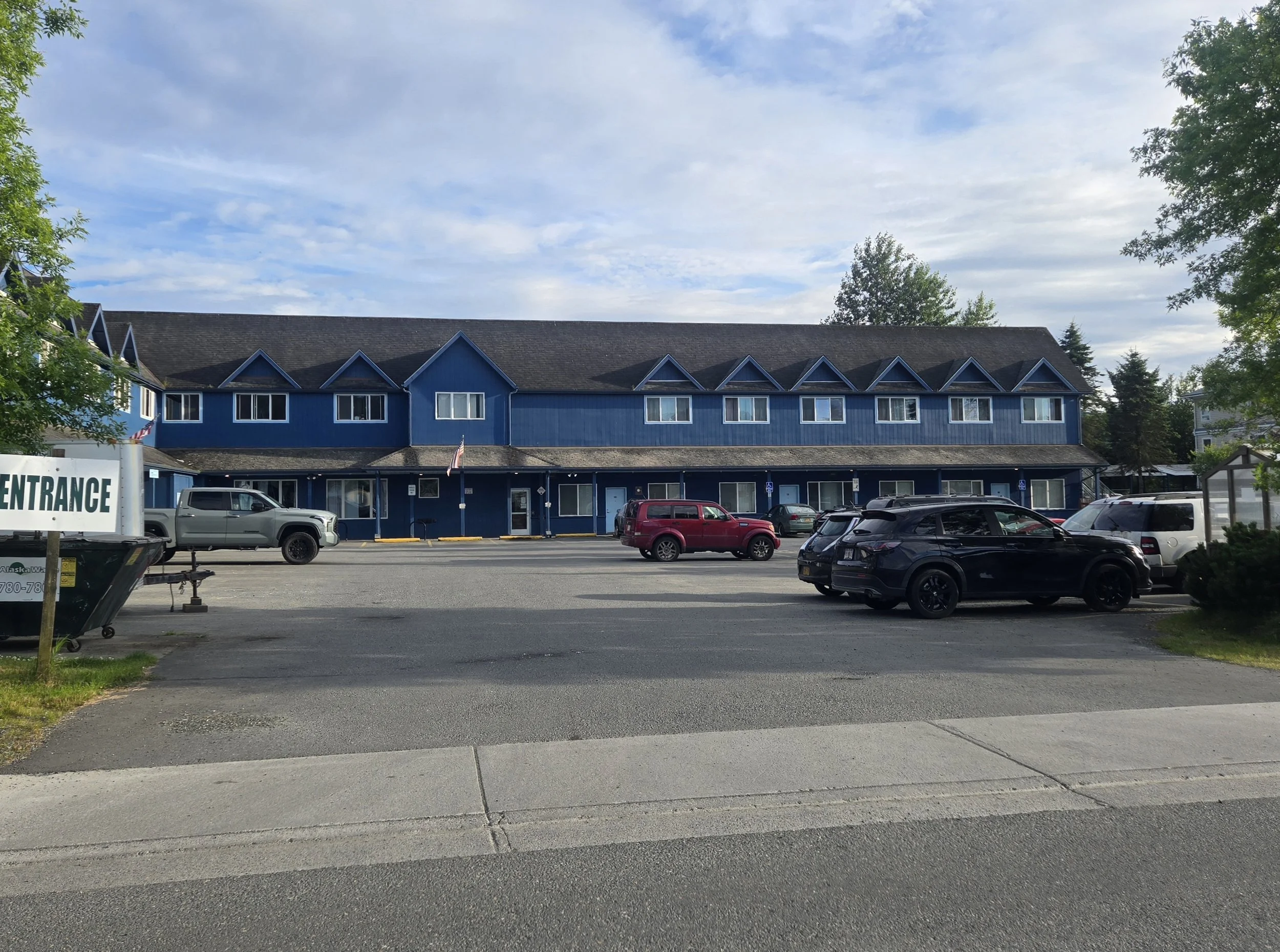A blue three-story building with dormer windows, parked cars in front, and a parking lot with trees and a partly cloudy sky in the background.
