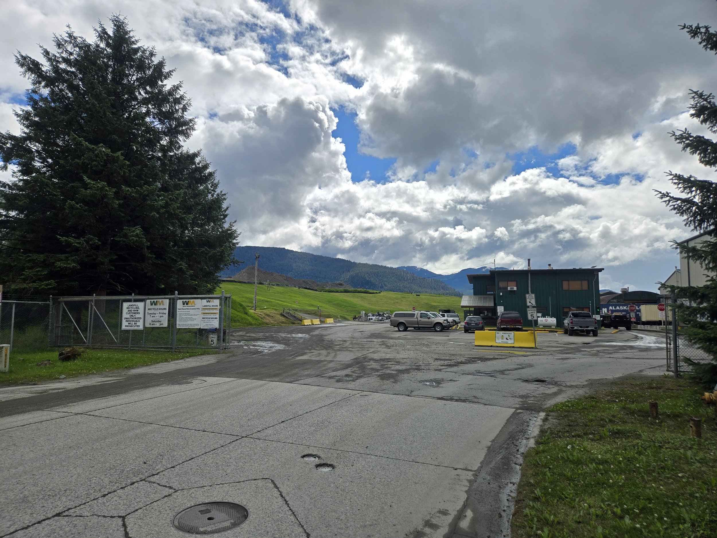 A parking lot with several parked vehicles and fencing with signs, adjacent to a grassy area with trees, and mountains in the background under partly cloudy sky.