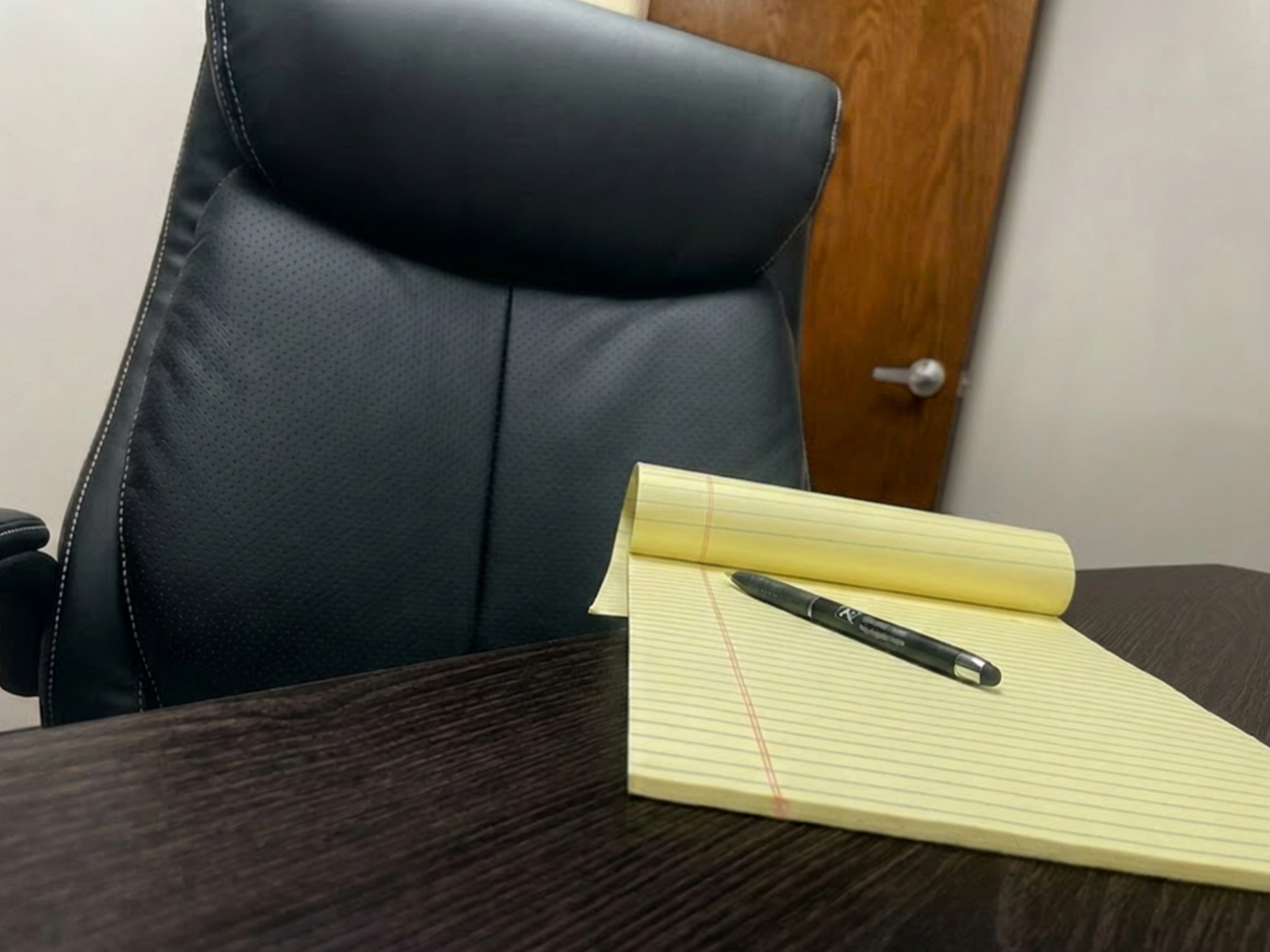 An office environment with a black office chair, a yellow notepad with red lines and a black Sharpie pen on top on a dark wooden desk. Behind the desk, a wooden door with a silver doorknob is visible, along with a plain white wall.