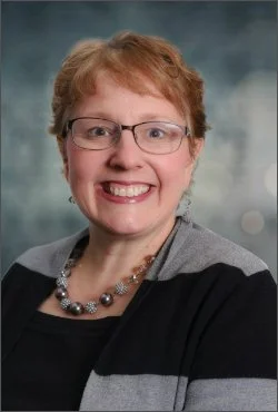 A smiling woman with short red hair, glasses, and a necklace, wearing a gray and black striped top, against a blurred background.