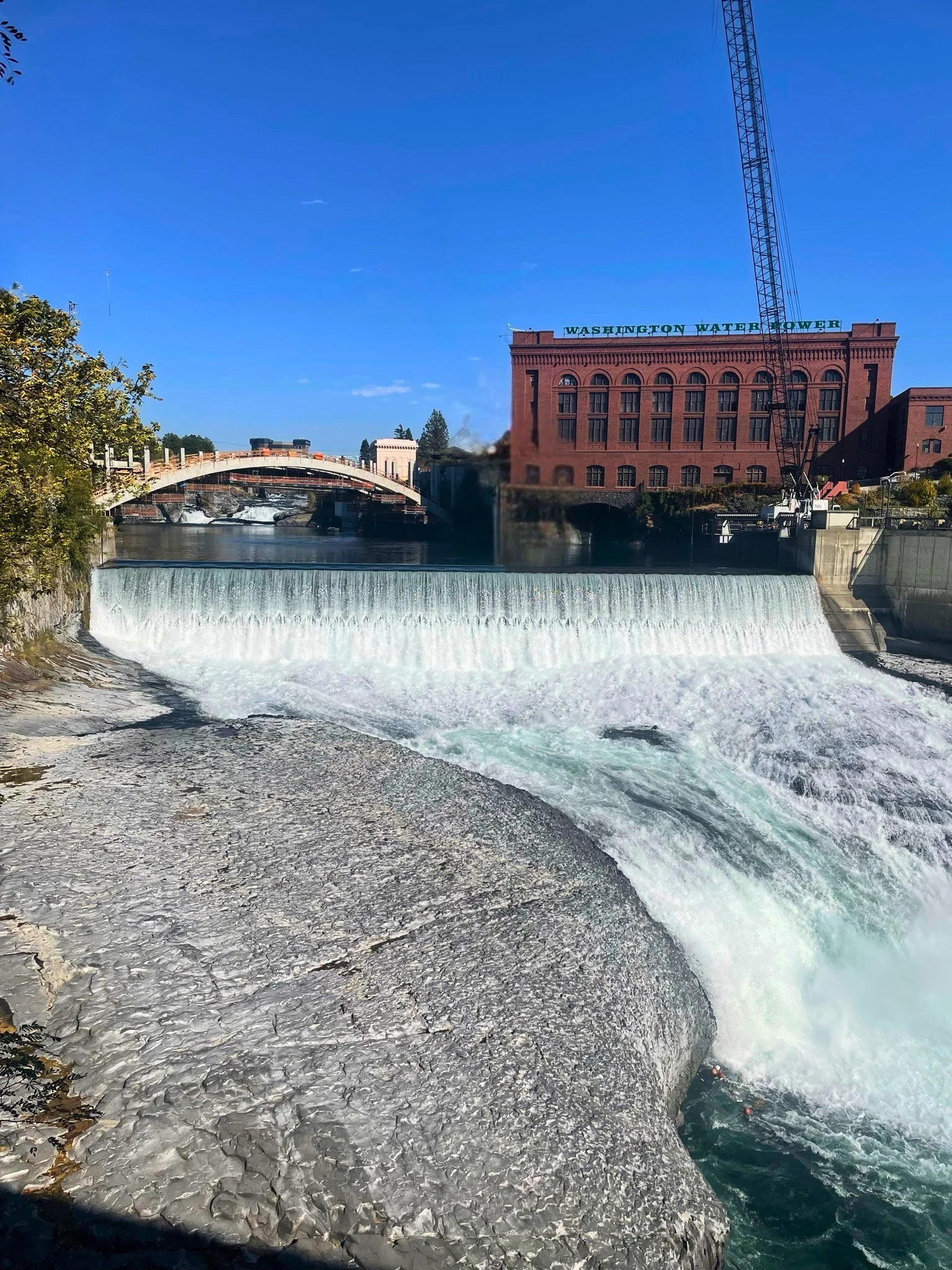 View of a river with a small waterfall, a bridge, and a brick building with a sign reading 'Washington Water Power' under a clear blue sky.