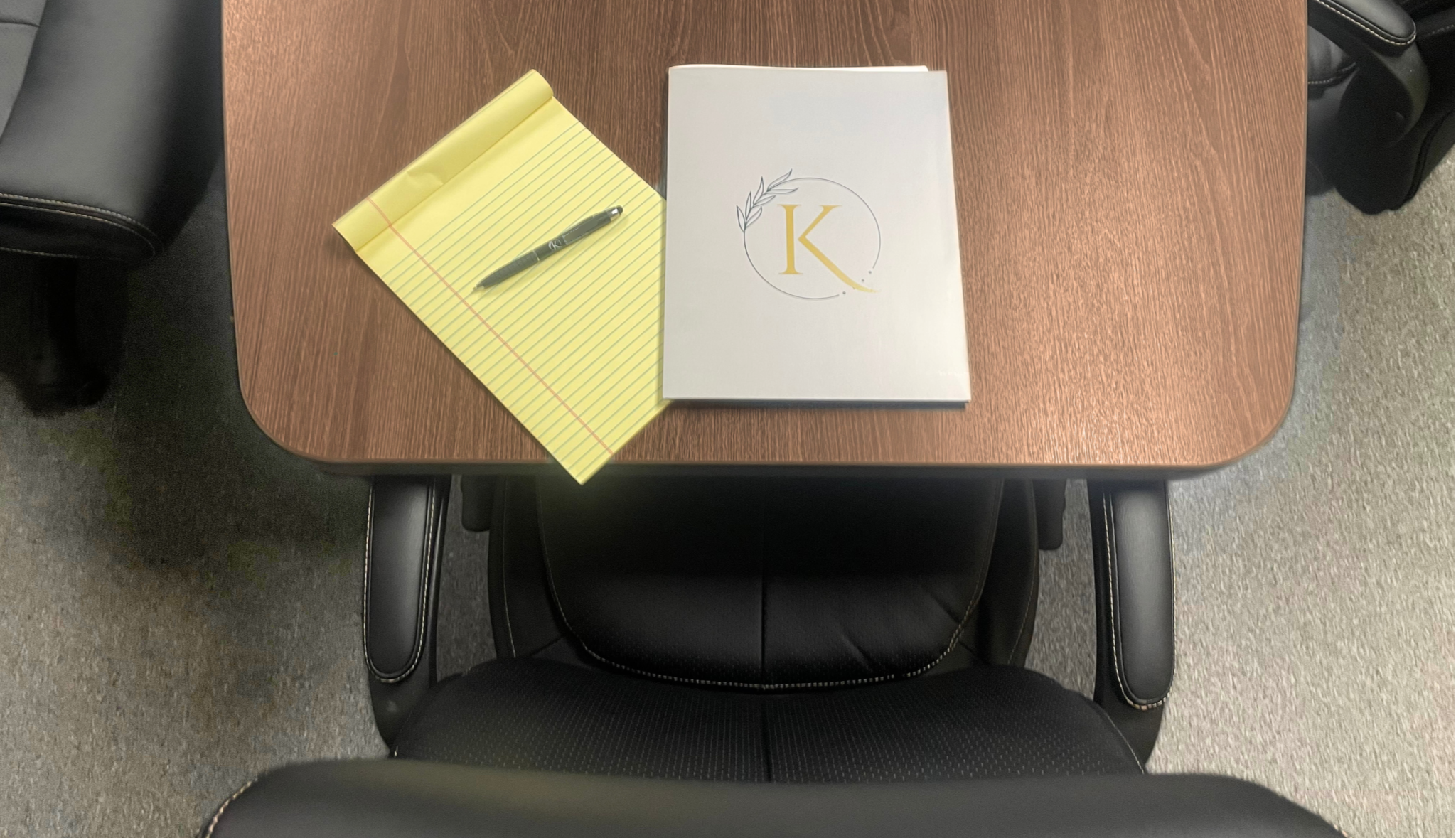 A wooden conference table with a black leather office chair. On the table are a black pen on a yellow lined notepad and a white book with a gold and gray logo of a letter 'K' surrounded by a circle and leaves.