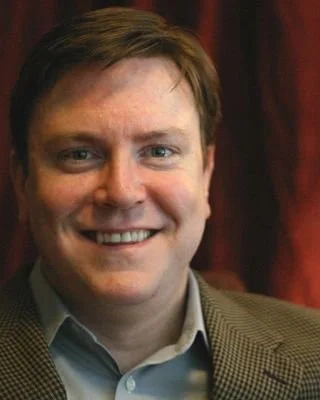 A smiling man with brown hair, wearing a light dress shirt and brown blazer, standing in front of a red curtain.