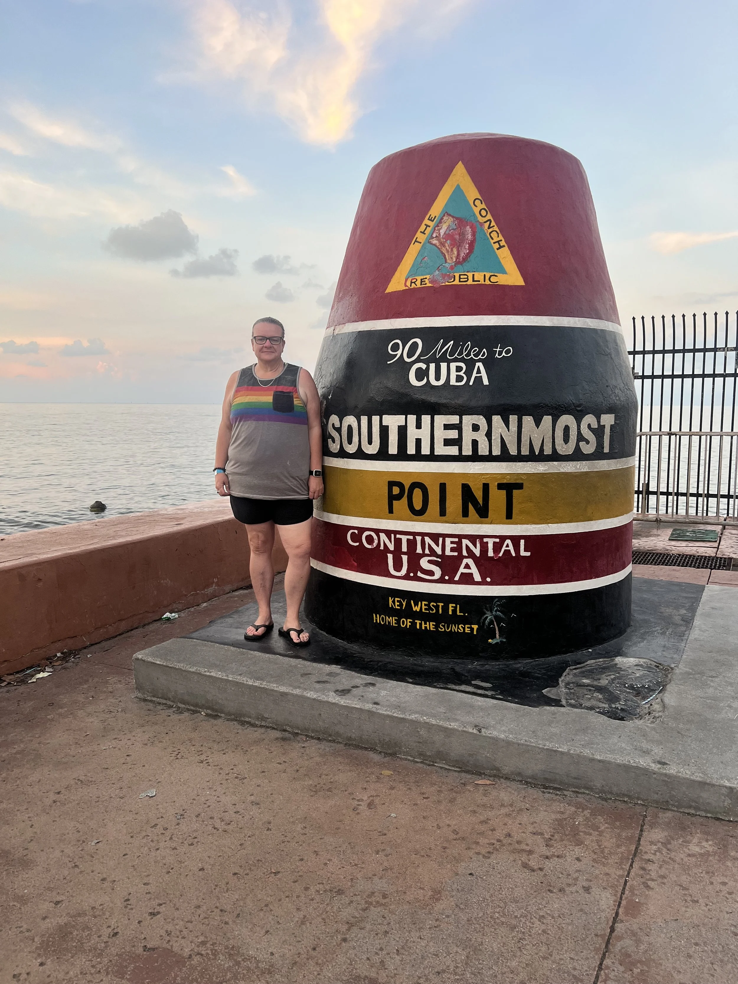 A woman wearing glasses and a colorful striped tank top standing next to a large, painted border marker indicating the southernmost point of the continental U.S. in Key West, Florida, with a sunset sky and water in the background.