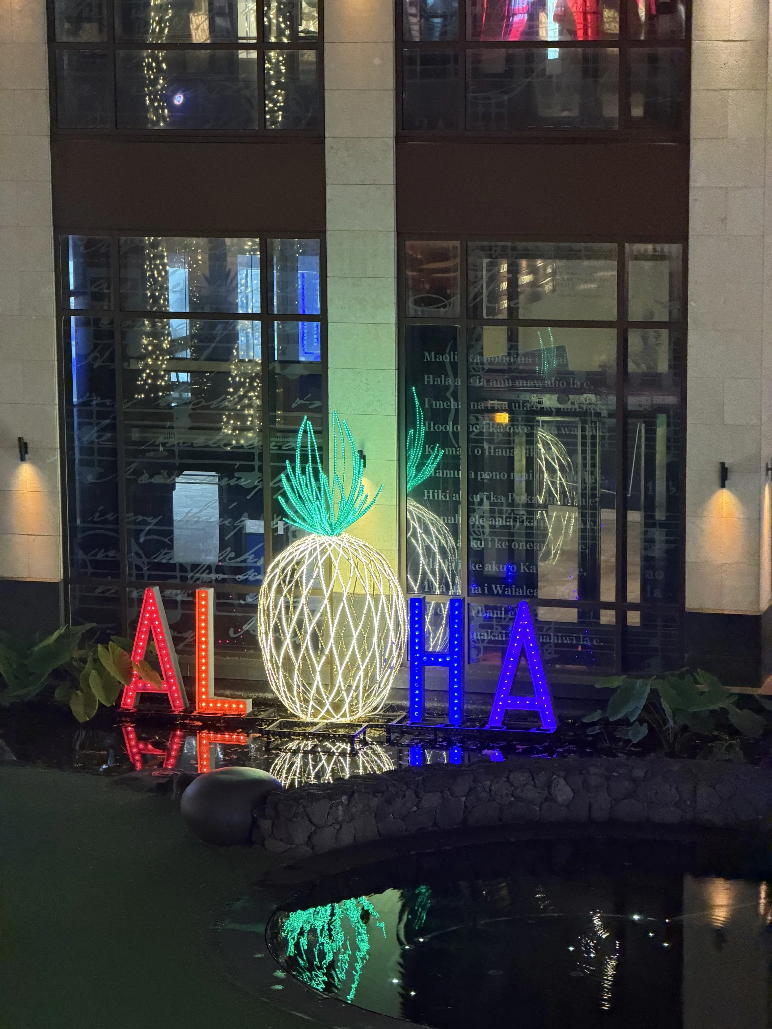 Nighttime scene of illuminated holiday decorations in front of a glass building, including a large pineapple-shaped light display and the word 'ALOHA' spelled out with colorful lights, which are reflected in a nearby pond.
