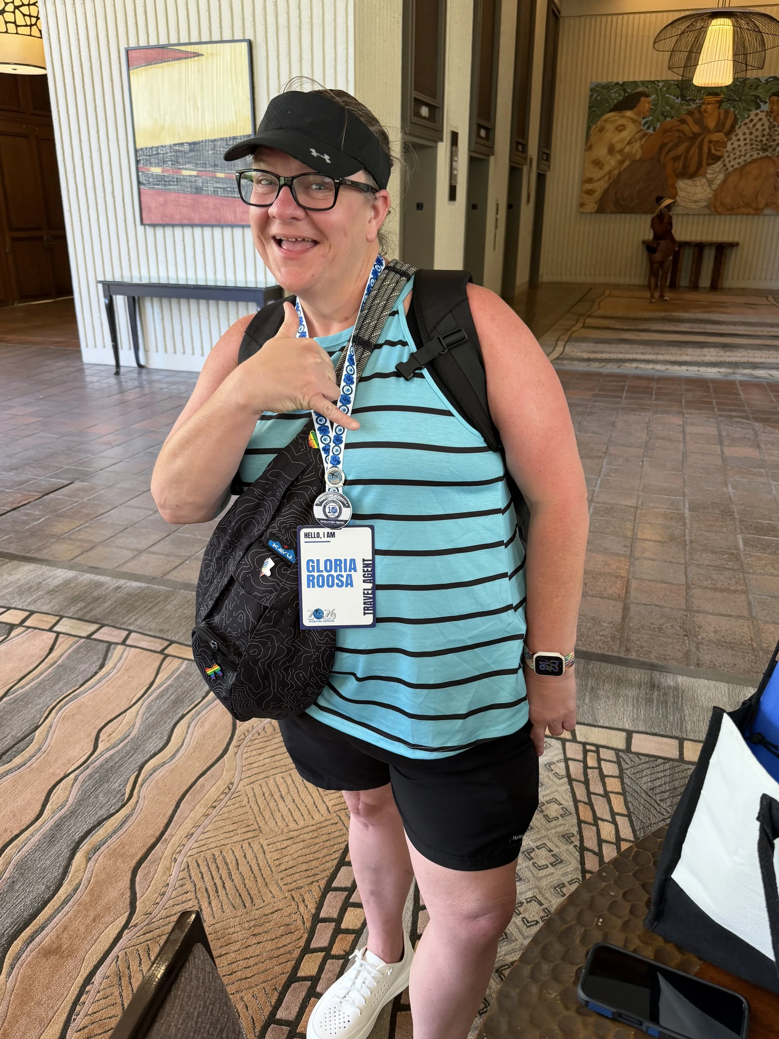 A smiling woman wearing glasses, a black visor, and a turquoise striped tank top, giving a thumbs-up gesture, standing in a hotel lobby with artwork on the walls and a patterned carpet.