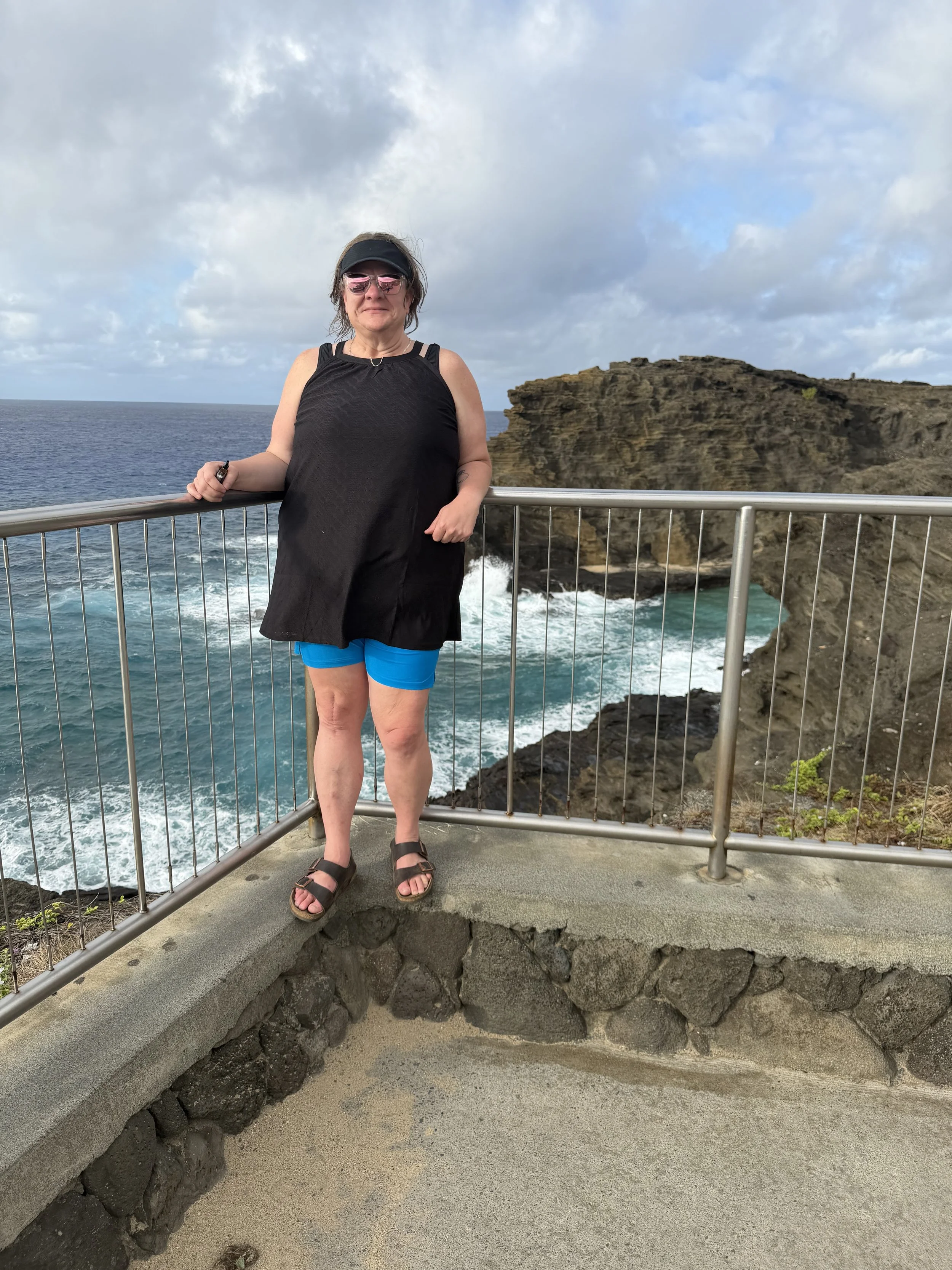 A woman standing on a viewing platform near a rocky coastline, wearing sunglasses, a black sleeveless top, blue shorts, and sandals, with the ocean and cloudy sky in the background.