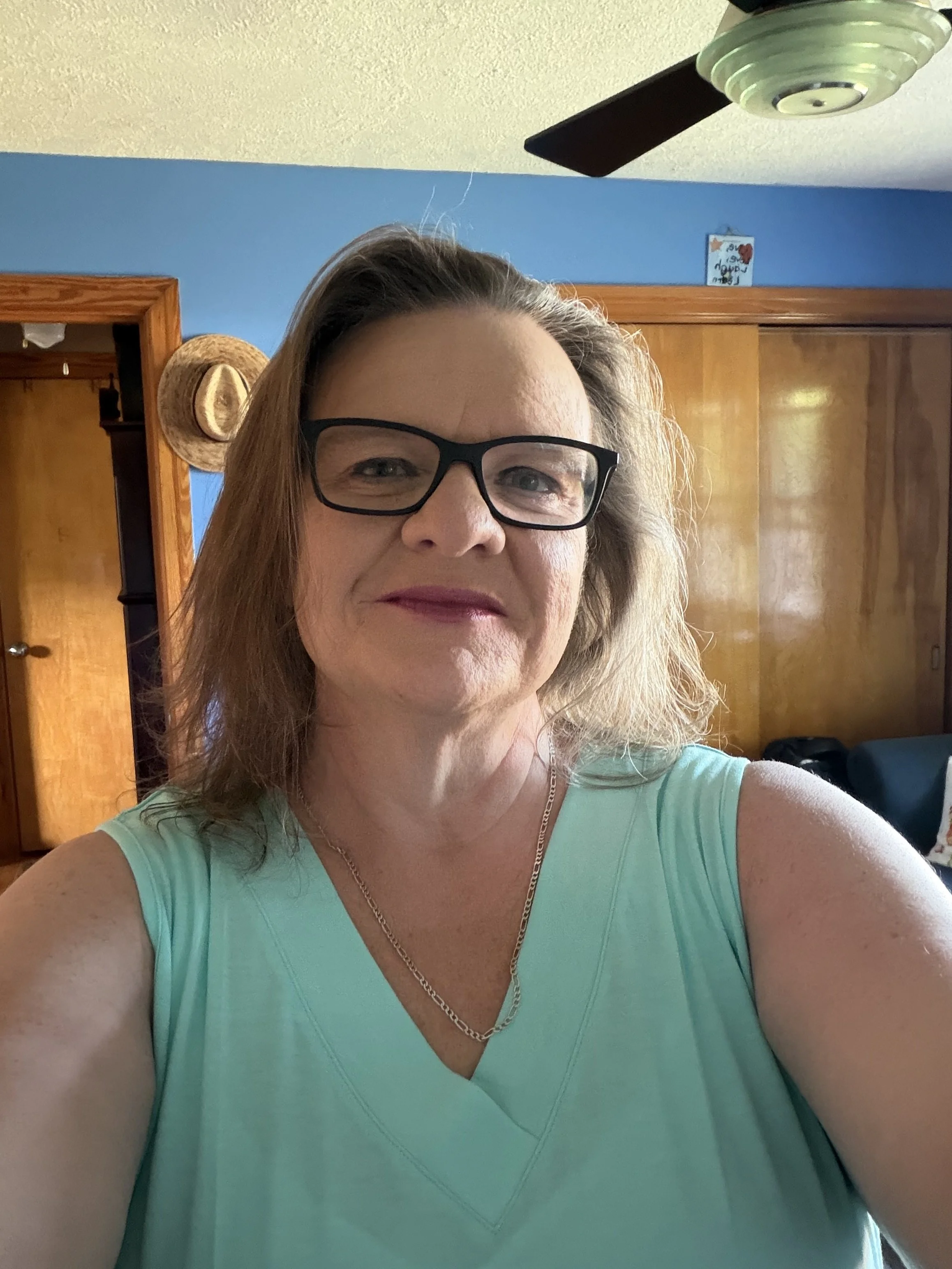 A woman with blonde hair, glasses, and a light blue sleeveless top taking a selfie in a room with blue walls, a wooden door, a wall hanging, and a ceiling fan.