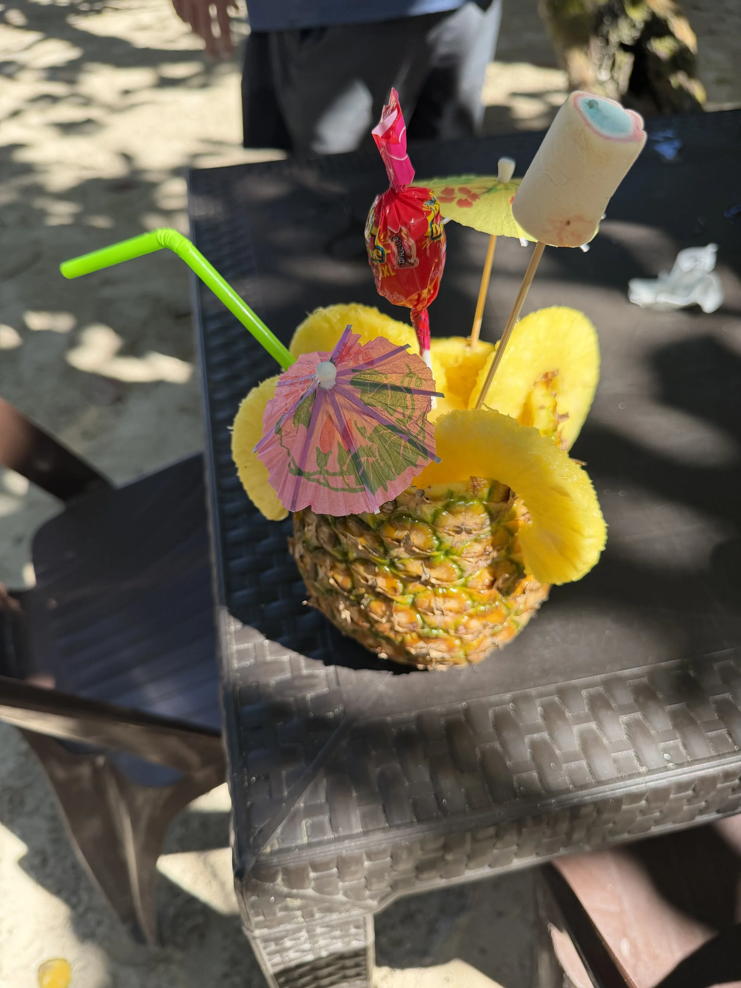 A pineapple is decorated as a drink with colorful accessories, including paper umbrellas, a lollipop, a paper pineapple, a small pink umbrella, and a neon green straw, set on a black metal table outdoors.