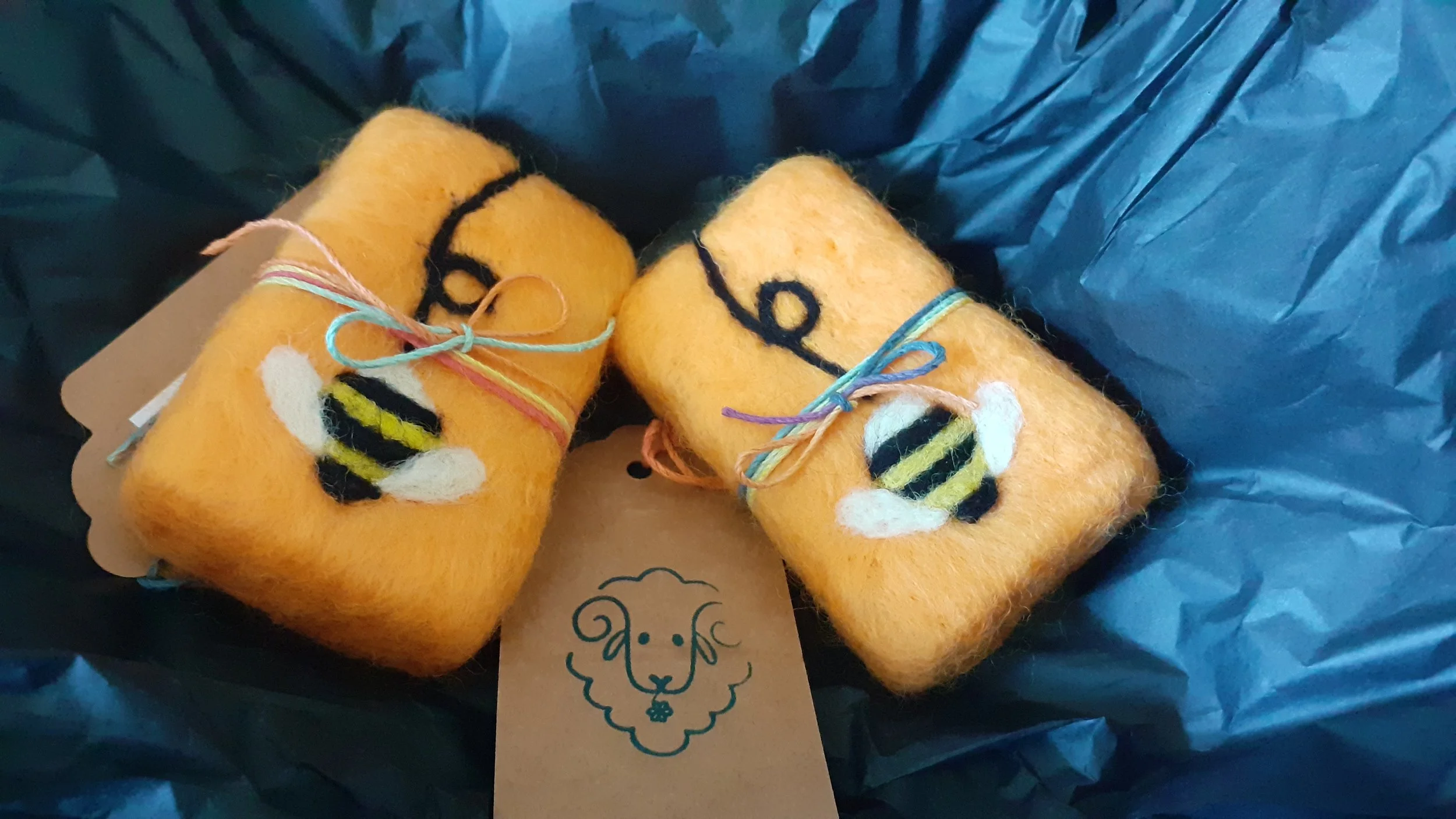 Two yellow felted soaps decorated with black and white yarn patterns, tied with colorful strings, placed on a blue surface with a brown tag and a lion illustration.