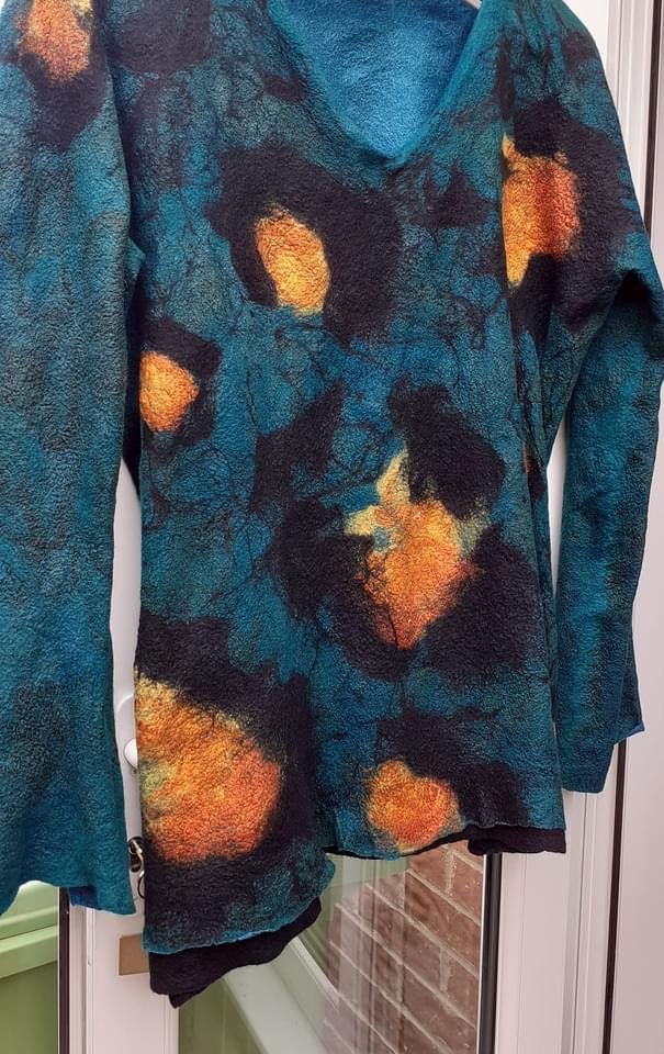 Close-up of a multicolored felted jacket with blue, black, orange, and yellow abstract patterns hanging near a window.
