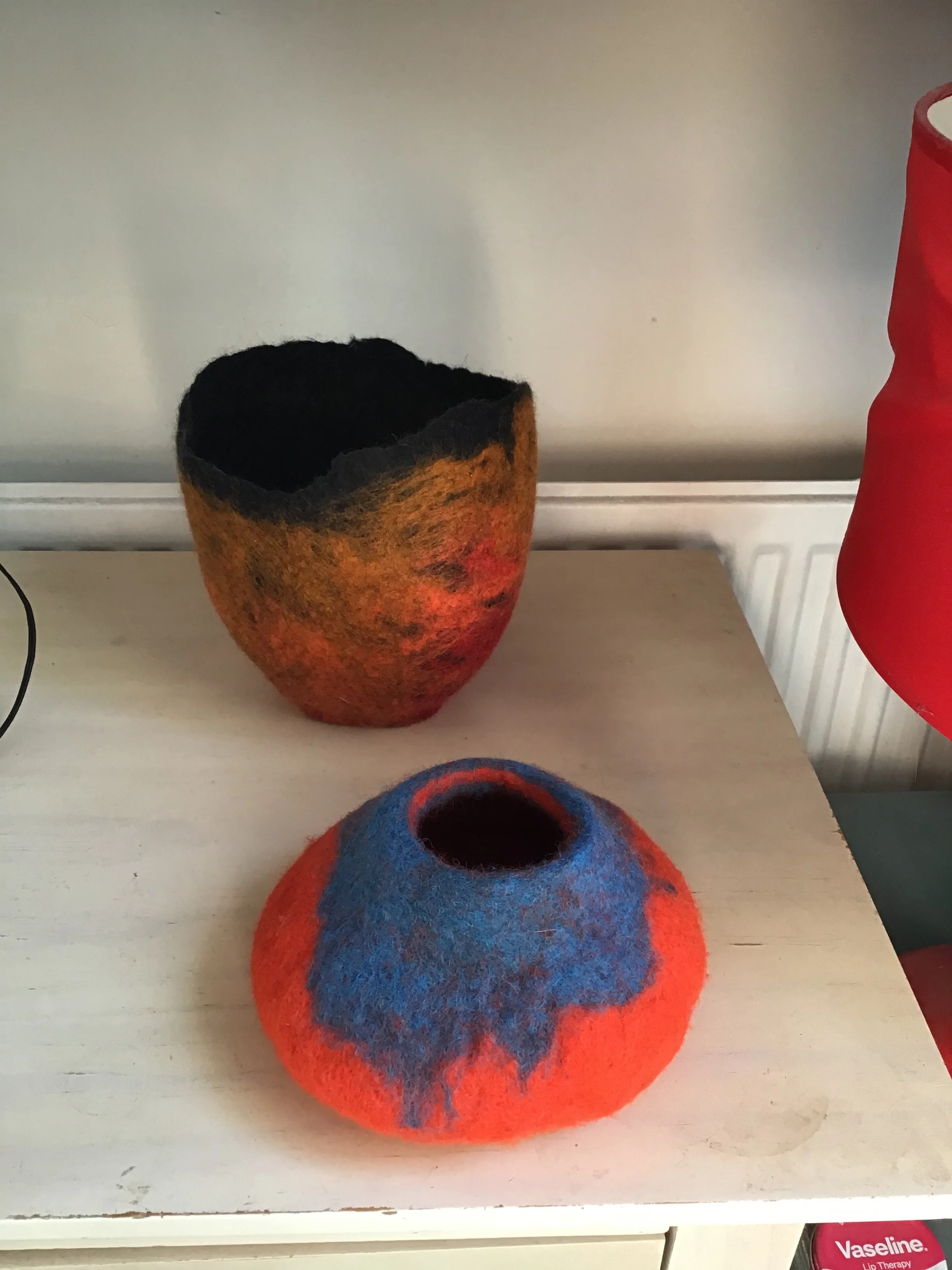 Two handmade felted wool bowls with vibrant colors, one in shades of black and orange, and the other in blue and orange, placed on a white surface.