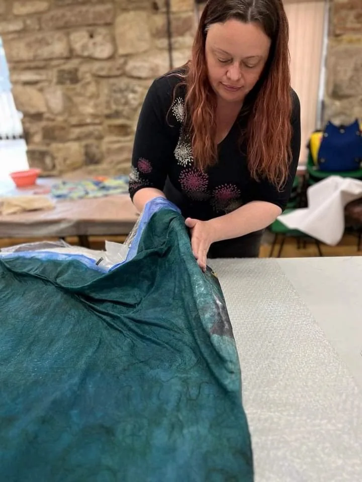 A woman with long reddish-brown hair, wearing a black shirt with colorful patterns, is unfolding or inspecting a large green fabric or material on a table in a room with brick walls.