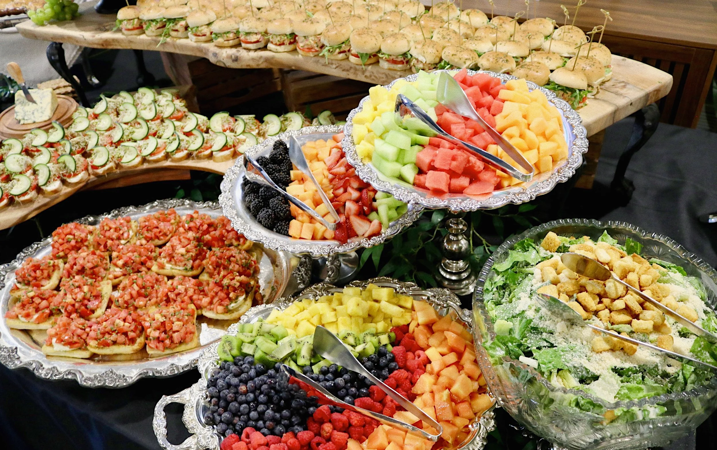 Appetizer table featuring smoked chicken salad sandwiches, cucumber sandwiches, assorted fruits, Caesar salad, and bruschetta, prepared by Silver Platter Catering