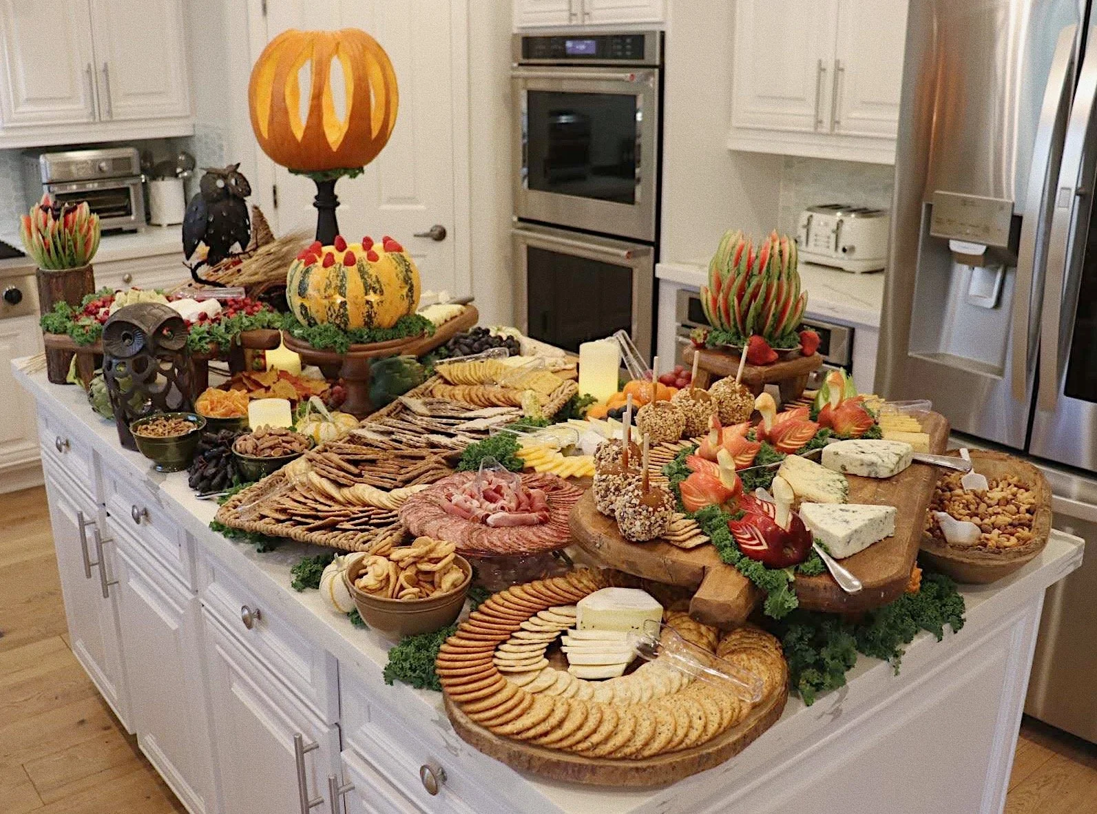 A Halloween-themed appetizer and snack table featuring assorted cheeses, crackers, meats, artistically carved watermelons and pumpkins, and delicate carved apple ducks, prepared by Silver Platter Catering