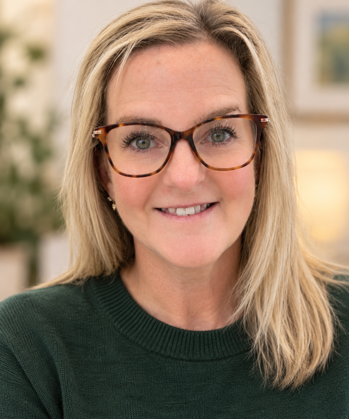 Close-up photo of an Anxiety Therapist with blonde hair, wearing tortoiseshell glasses and a dark green sweater, smiling at the camera.