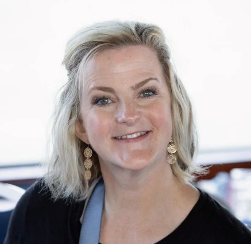 A woman with blonde, shoulder-length hair, smiling at the camera, wearing gold earrings and a black top.