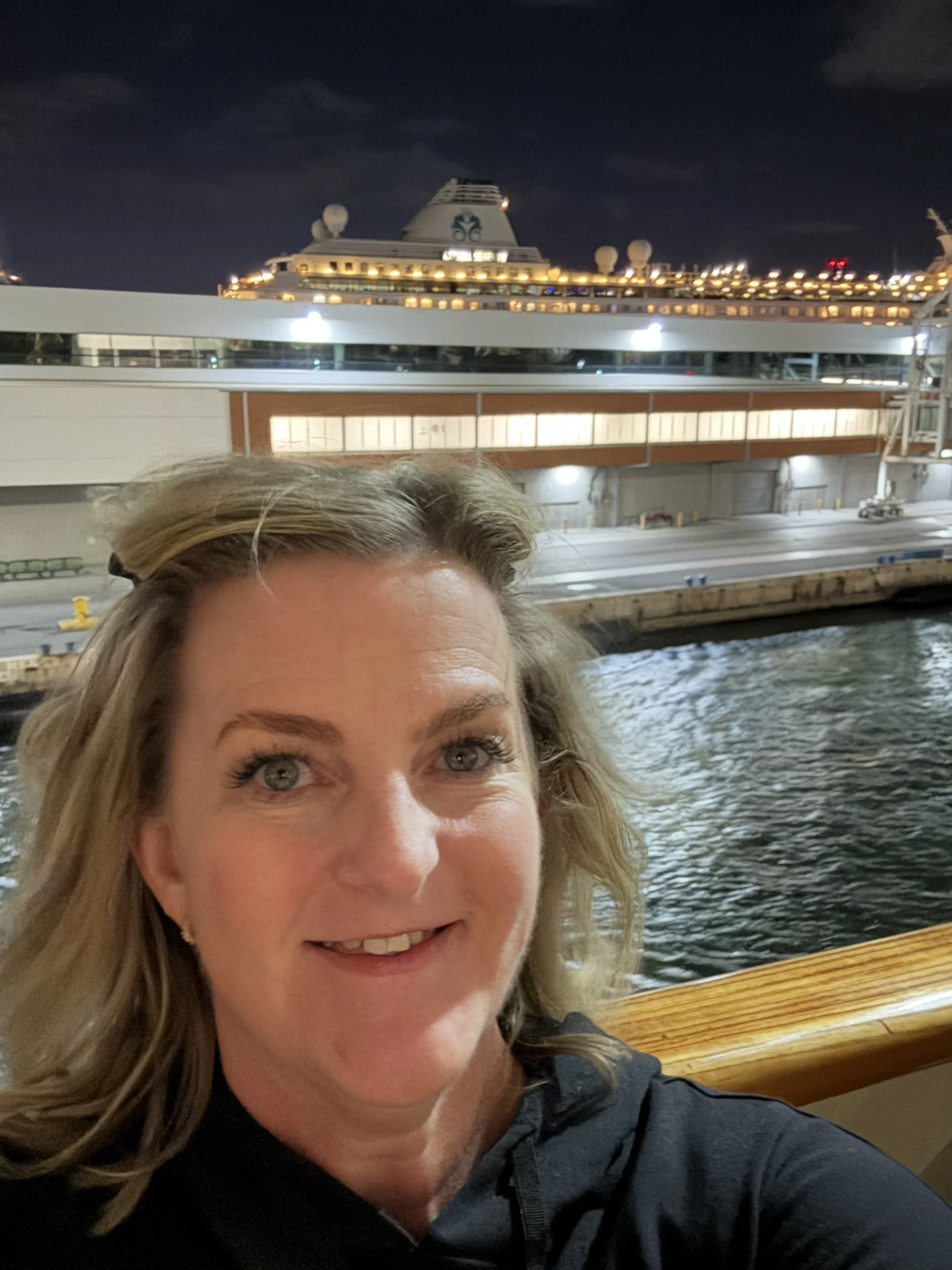 A woman with blonde hair and blue eyes smiling on a boat at night, with a large cruise ship illuminated in the background.
