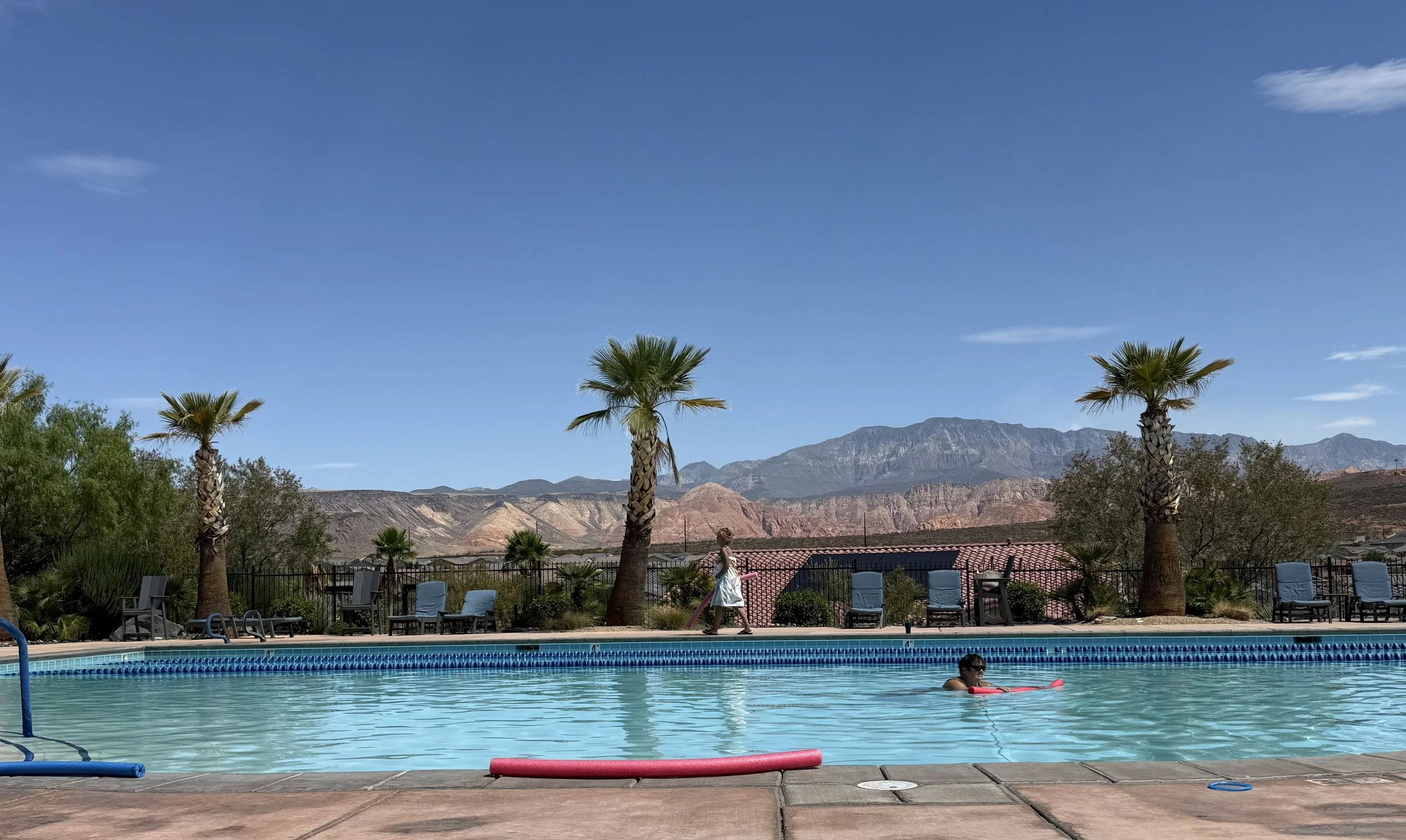 Outdoor swimming pool with a child swimming with a float, surrounded by lounge chairs, palm trees, and a mountain range in the background under a clear blue sky.