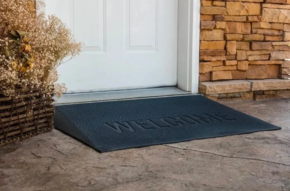 A black welcome doormat placed in front of a white house door, with a basket of small white flowers on the left and a brick wall on the right.