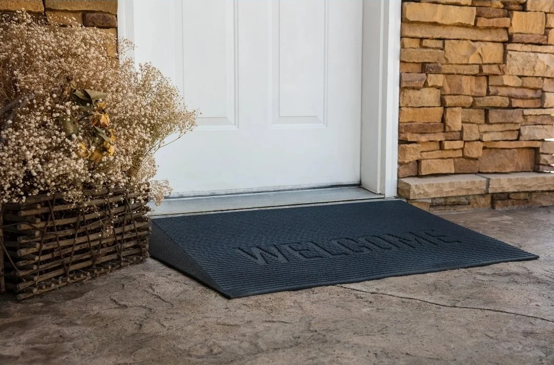 A black welcome mat in front of a white door, with a wicker basket containing dried flowers and a brick wall on the sides.