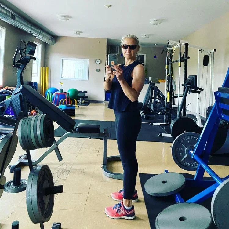 Woman taking a selfie in a gym, wearing sunglasses, black workout clothes, and pink sneakers, surrounded by gym equipment.