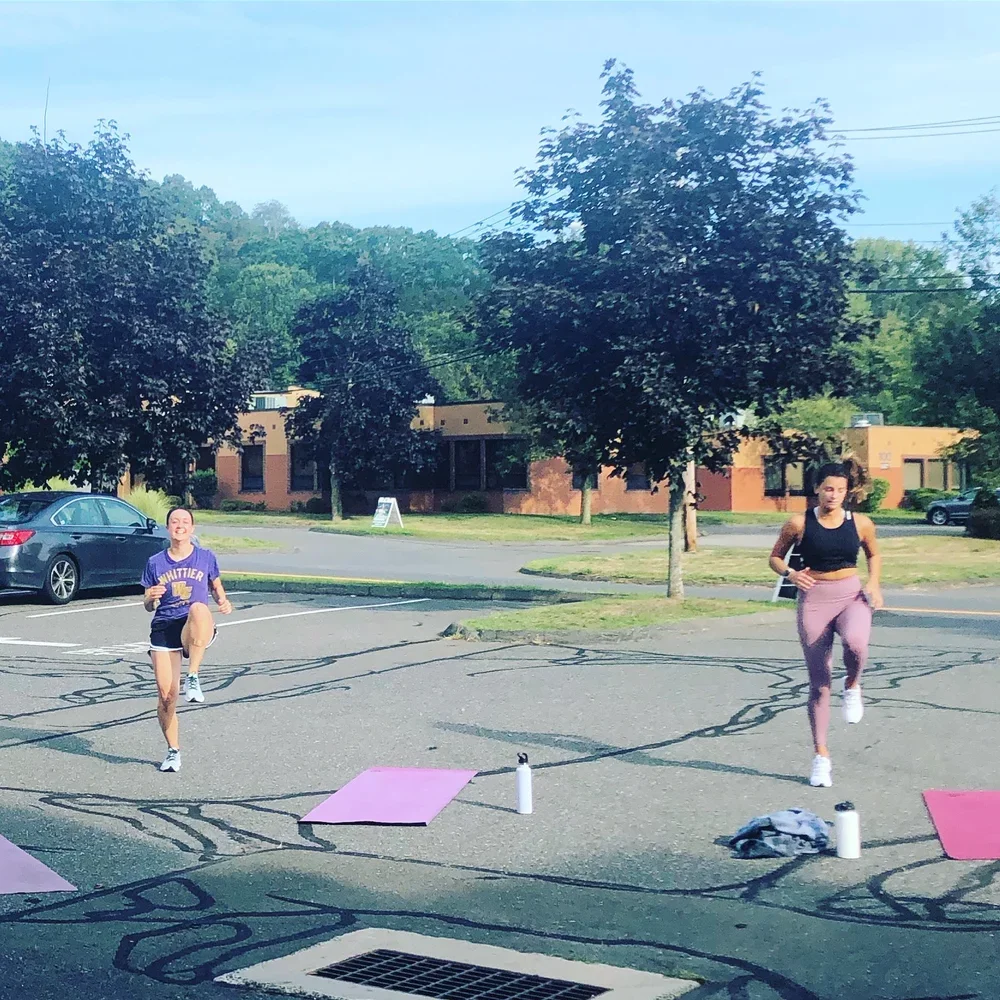 Two women are doing a workout in a parking lot with yoga mats, water bottles, and water bottles, under trees with a building and parked cars in the background.