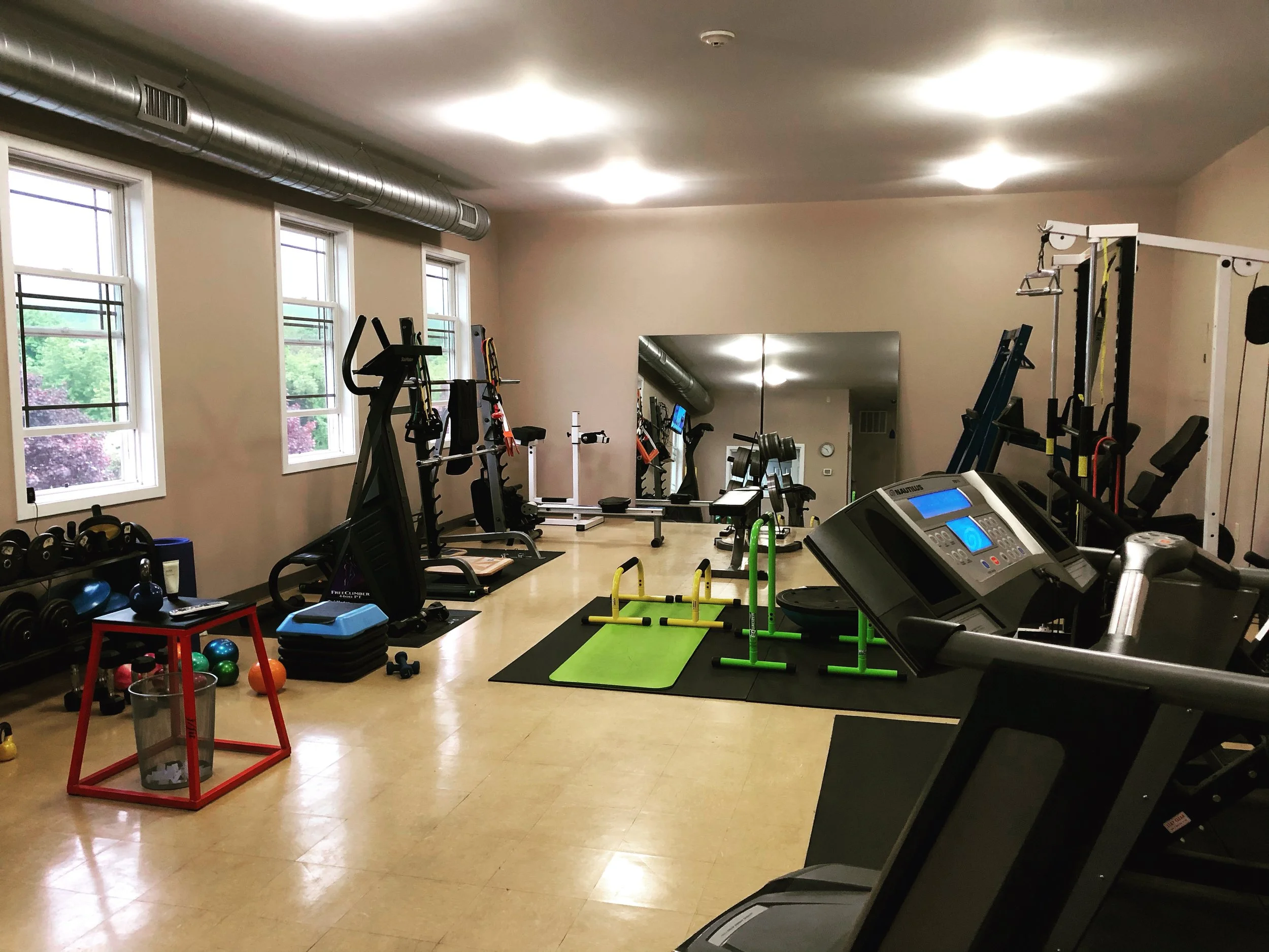 A gym room with various exercise equipment including treadmills, weight benches, and resistance machines. There are several windows with natural light and some fitness accessories like kettlebells and stability balls in the room.