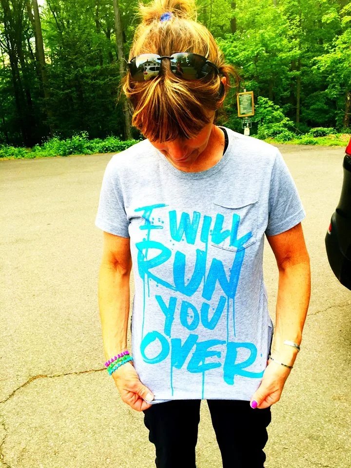 A woman standing outdoors in a parking lot, wearing sunglasses on her head, a light gray T-shirt with blue text that reads 'I WILL RUN YOU OVER,' black pants, and colorful bracelets on her left wrist. She is looking down.
