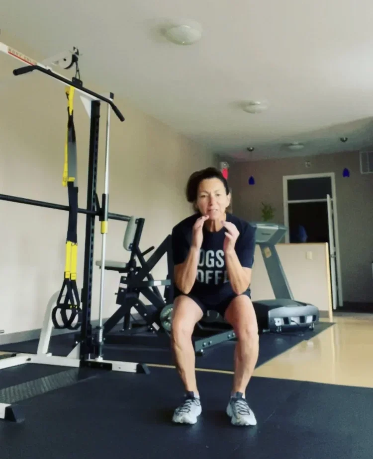 An elderly woman in black athletic clothing sits on a workout bench in a home gym, holding a yellow dumbbell with her hands near her face. The gym has various exercise equipment, including a squat rack, treadmill, and additional weights.