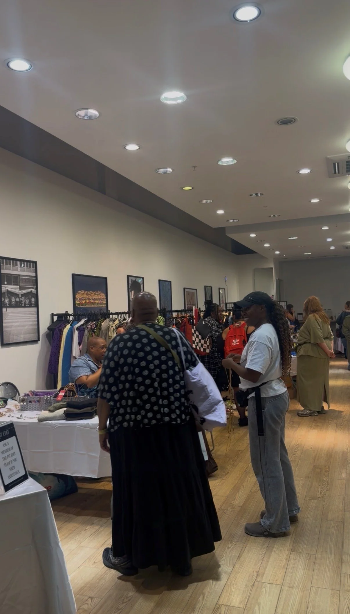 People browsing clothing and accessories at an indoor market or event with framed pictures on the wall and ceiling lights.