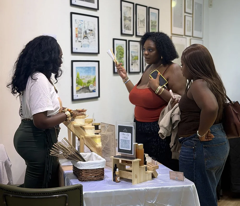 Three women at an art fair or gallery looking at skincare or beauty products on a vendor table.
