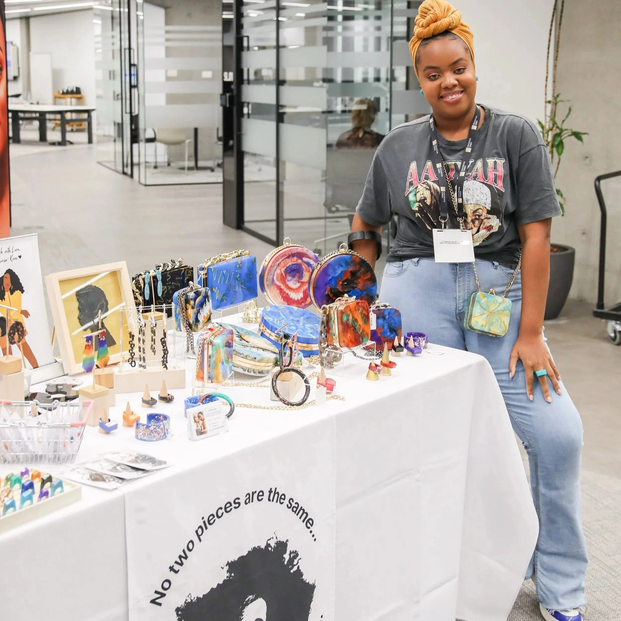 A young woman with a head wrap and earrings standing behind a table displaying colorful jewelry and accessories at an indoor event or market. She is smiling and wearing a graphic t-shirt and jeans. The background shows glass doors and a modern office or gallery space.