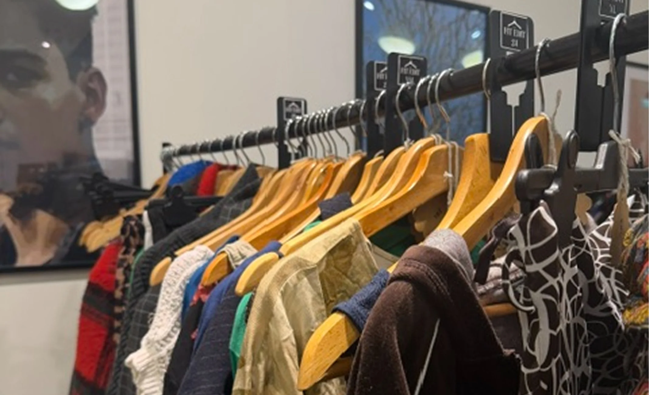 Clothing rack with various colorful shirts and jackets hanging on wooden hangers,with a man’s reflection in a mirror behind it.