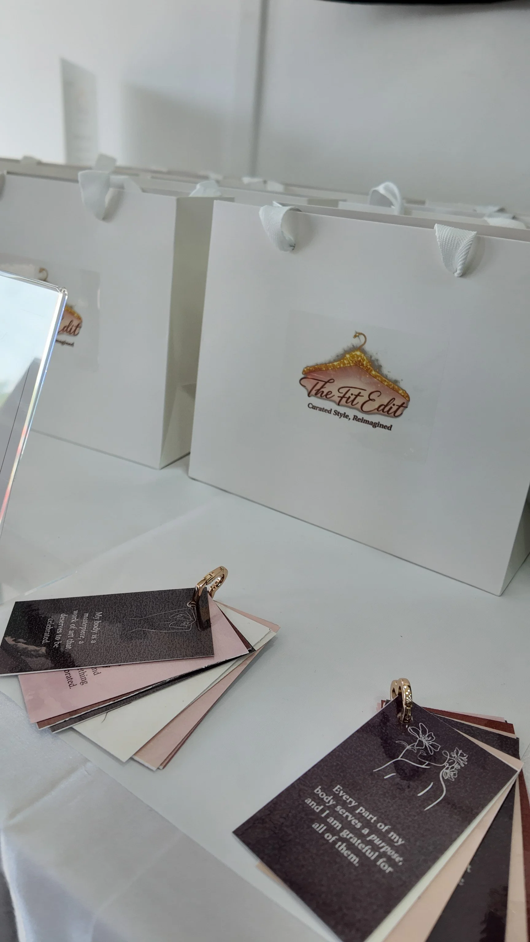 White display cards with black ornaments and text, some cards have the logo 'The Fit Edit' with a hanger and gold accents, placed on a white surface with small booklets, hooks, and a mirror reflecting part of the display.