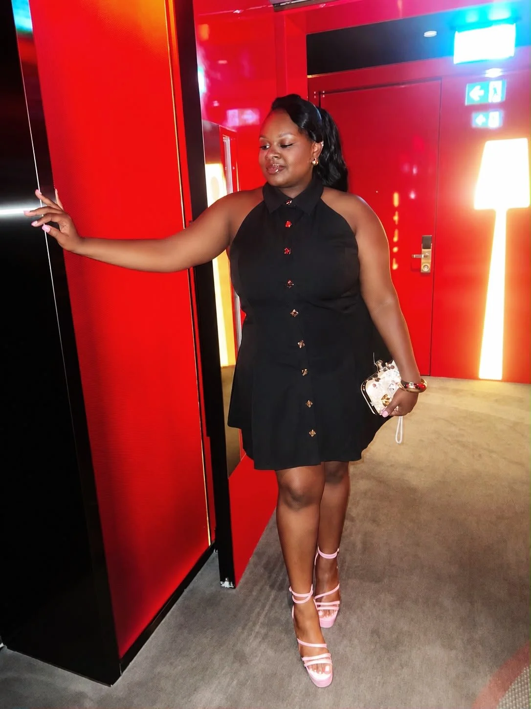 A woman in a black dress with pink heels standing in front of a red wall, holding a small white purse with a chain strap.