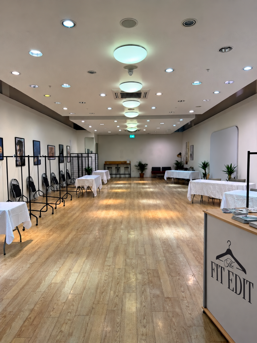 Empty event or meeting room with white tablecloth-covered tables, chairs, framed photos on the walls, and potted plants, set up for a function or gathering.