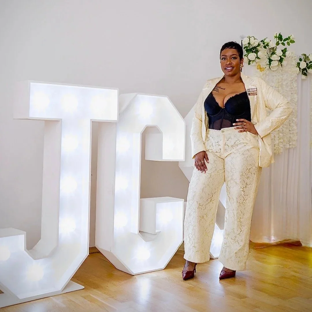 woman standing next to illuminated white letter signs that spell 'JAC', in a light-colored suit and black top, with white flowers in the background.