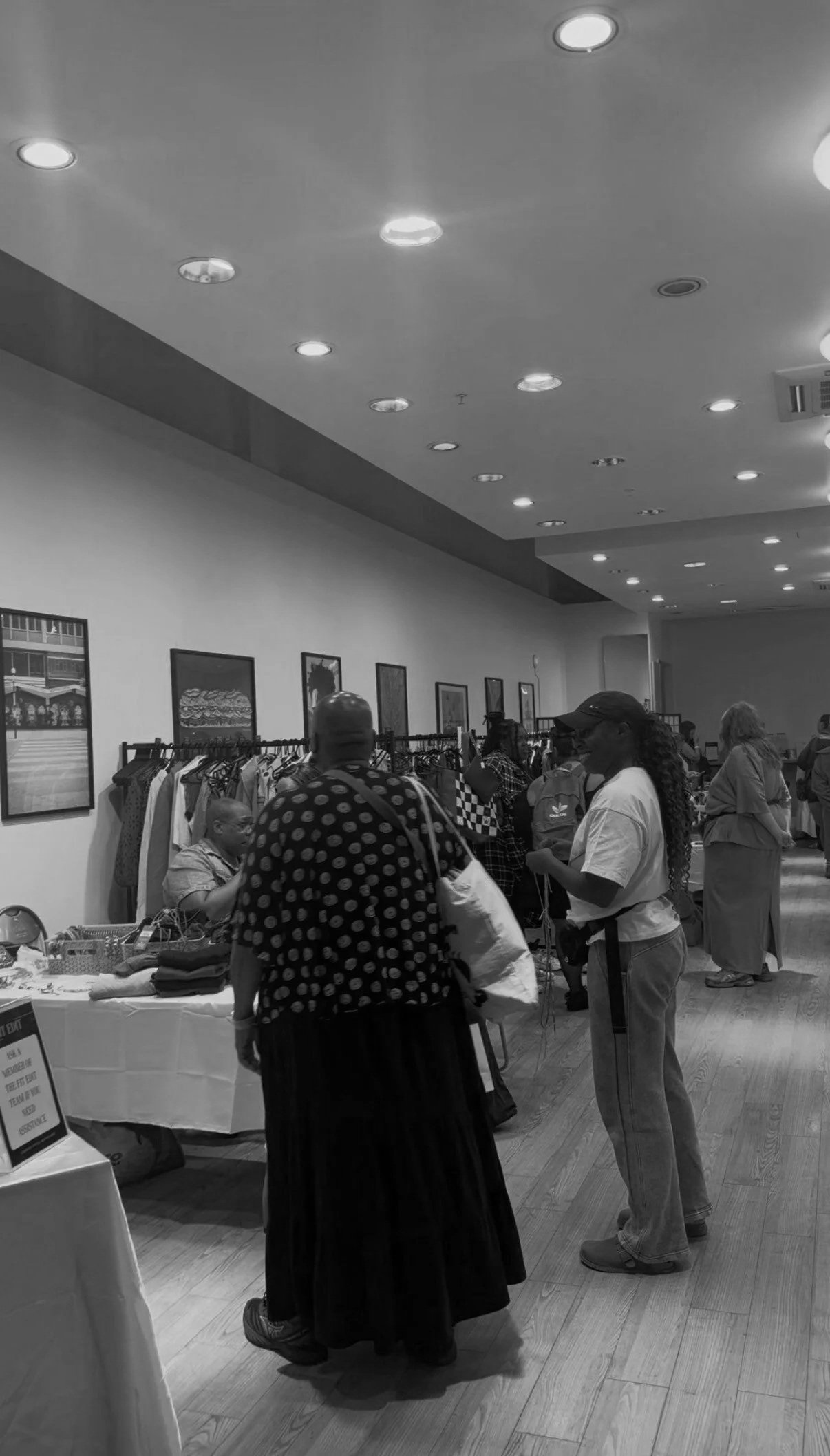 People shopping at an indoor market or event with clothing racks and tables with items for sale in a well-lit room.