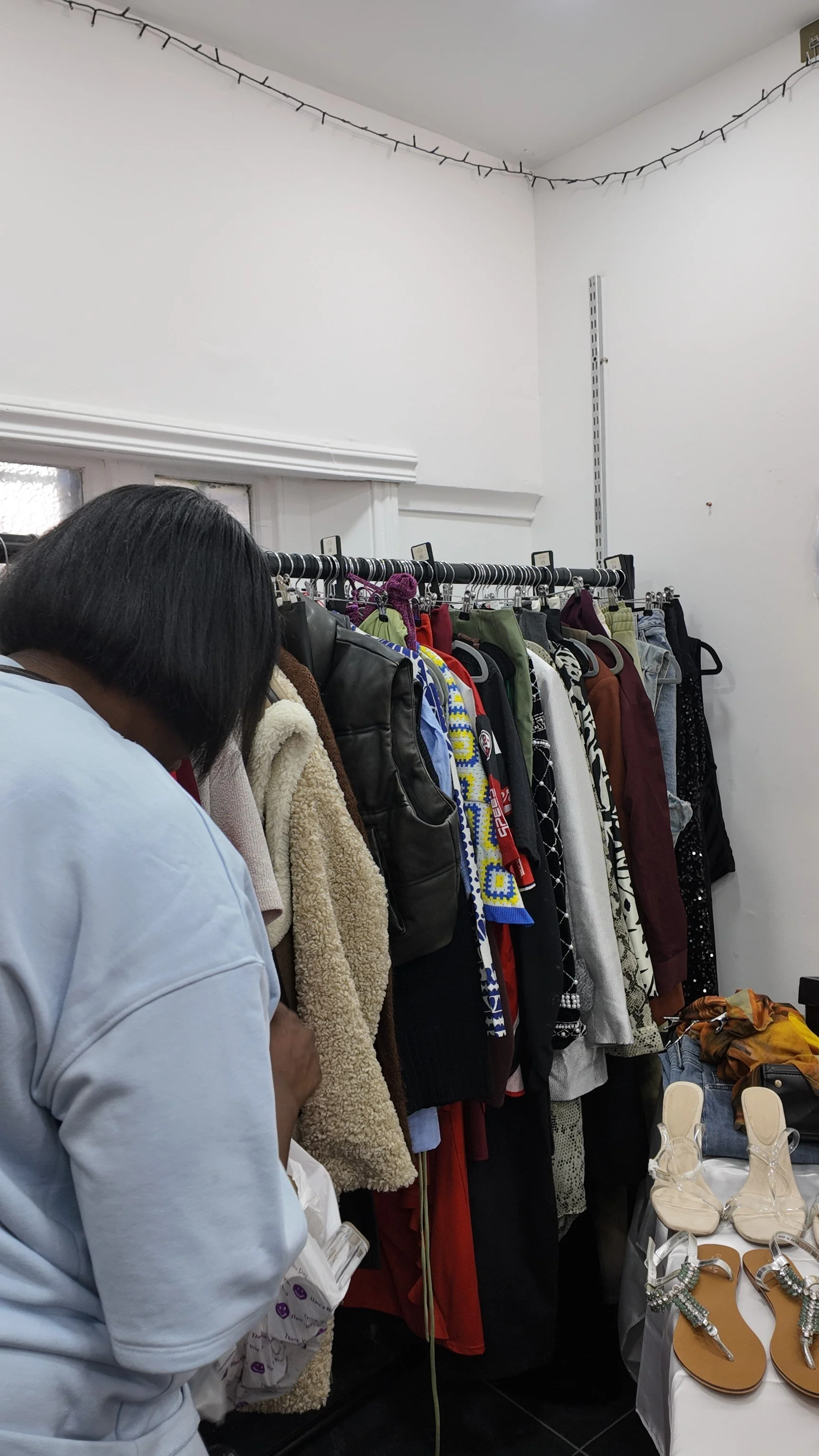 A person browsing clothing and accessories at a thrift or vintage store. The clothing rack holds various jackets, sweaters, and dresses in different colors and styles. There is a table with sandals and jewelry displayed to the right.