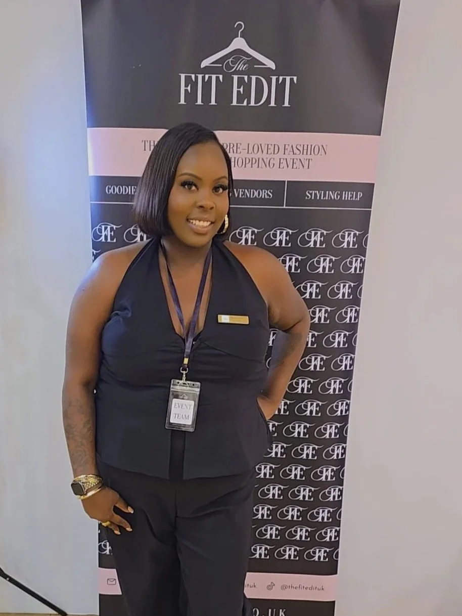 A woman standing in front of a banner at an event. She is smiling, wearing a black outfit with a name tag and an event badge. The banner behind her reads 'The Fit Edit' and mentions a pre-loved fashion shopping event.