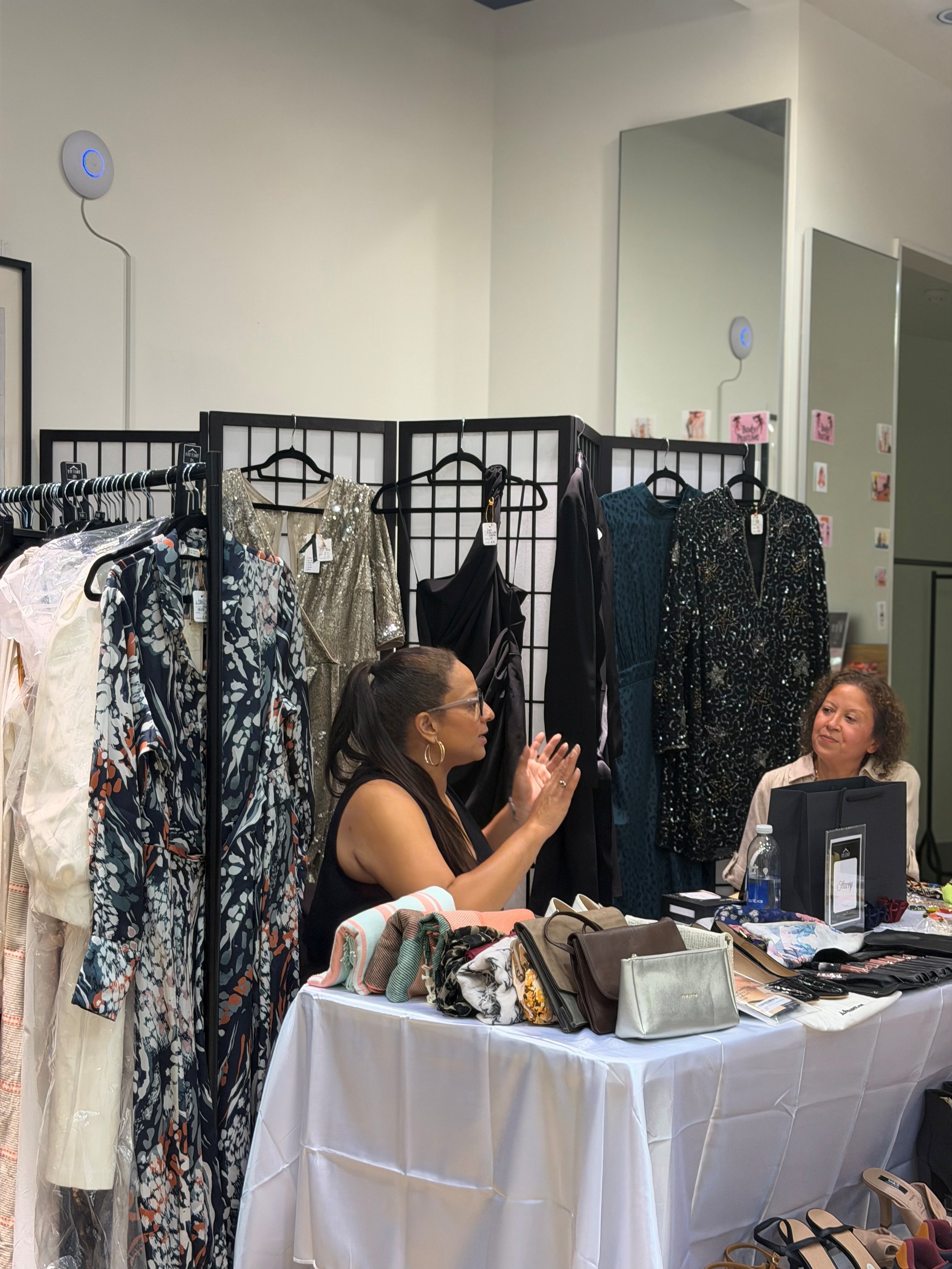 A clothing and accessories booth at a market or store, with dresses hanging on racks and handbags, scarves, and other accessories displayed on a table in front. Two women are engaged in conversation behind the table.