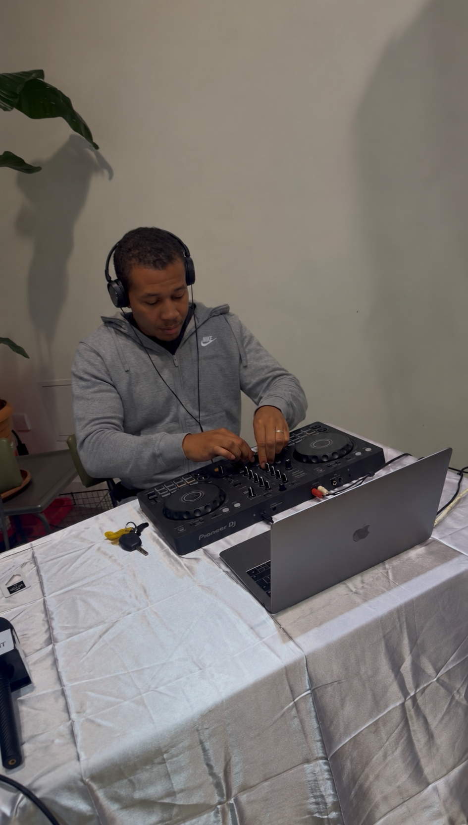A man DJing with headphones, using a Pioneer DJ controller, a laptop, and mixing equipment on a table covered with a white cloth.